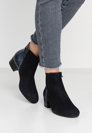 Bottines en daim noir avec talon et arrière à texture peau de serpent bleue, portées avec un jean skinny gris à ourlet effiloché sur un sol blanc.