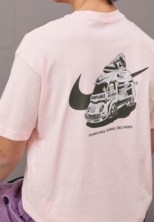 Person trägt ein hellrosa Shirt mit einem schwarzen Nike-Swoosh und einem Cartoon-Dim-Sum-Lieferwagen, der einen Turnschuh auf dem Dach transportiert.