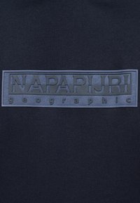 Σκούρο μπλε ύφασμα με ορθογώνιο κέντημα που εμφανίζει τη λέξη "NAPAPIJRI" με μεγάλα γράμματα και τη λέξη "geographic" με μικρότερα γράμματα από κάτω.