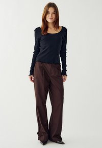 Čierny rebrovaný top s dlhými rukávmi a rozstrapkaným lemom, v kombinácii s voľnými hnedými nohavicami so šitými skladmi a širokými nohavicami, nosený s špicatými topánkami.