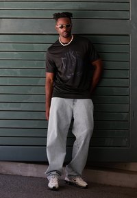 Must värvusega lühikeste varrukatega Nike T-särk, avarad helesinised teksad, valged ja mustad sneakerid ning kreemivärvi kaelakee rohelise tausta ees.