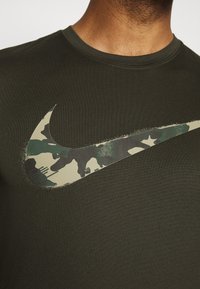 Camiseta verde oscuro con un gran logotipo de Nike con patrón de camuflaje impreso en el frente. El material es suave con un diseño clásico de cuello redondo.