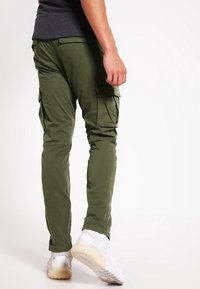 Oliwkowe spodnie bojówki z wieloma bocznymi kieszeniami, zwężane w nogawkach, wykonane z lekkiego materiału, zestawione z białymi sneakersami.