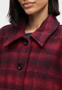 Red and black plaid wool coat with a collared neckline, black buttons, and a textured fabric. Close-up view of the front.