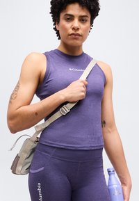 Personne portant un haut sans manches violet Columbia et des leggings, tenant une sangle beige clair d'une main et une bouteille d'eau violette dans l'autre.