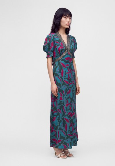 Femme portant une robe longue fleurie turquoise et magenta avec des manches bouffantes et une bordure en dentelle, debout devant un fond clair uni.