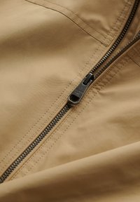 Close-up van een gedeeltelijk open metalen rits op beige stof met zichtbare stiksels.