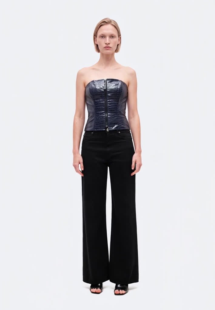 Un mannequin se tient devant un fond clair, vêtu d’un corset foncé sans bretelles, d’un pantalon noir à jambes larges et de sandales noires.