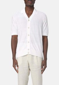 Camicia bianca a maniche corte con texture a coste verticali, colletto classico e patta con cinque bottoni. Indossata con pantaloni di lino chiari.