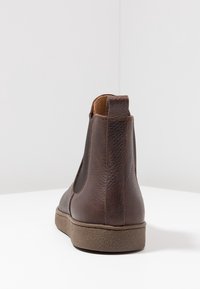 Botín de cuero marrón hasta el tobillo con una textura suave, paneles laterales elásticos y una suela de goma con un ligero tacón. Vista trasera mostrada.