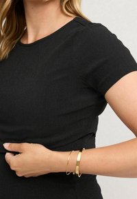 Zwart geribbeld kortmouwen shirt met een ronde halslijn. Een hand met twee gouden armbanden wordt getoond terwijl deze het taillegebied aanpast.