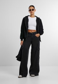 Felpa nera con zip sopra una maglietta bianca corta, abbinata a pantaloni cargo neri a gamba larga con tasche laterali e orli sfilacciati. Sneakers marroni con suola in gomma.