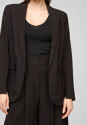 Kvinde iført en sort blazer, sort ærmeløs top og sorte højtaljede bukser mod en ensfarvet grå baggrund.