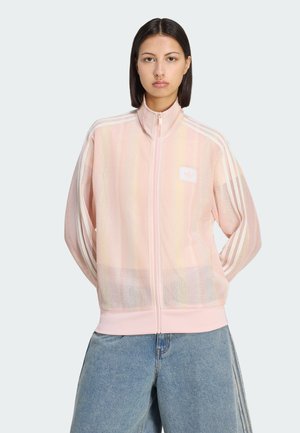 Junge Frau mit glatten dunklen Haaren trägt eine hellrosa Mesh-Zipjacke mit weißen Streifen und hellblaue weite Jeans, steht mit den Händen hinter dem Rücken.