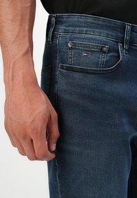 Lähivõte käest, mis puhkab tumedate siniste teksade kõrval, mille coin pocketil on väike Tommy Hilfiger logo.