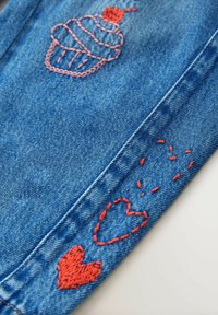 Tissu en denim avec broderie rouge représentant un cupcake et des formes de cœur. Le jean est de couleur bleu clair avec des détails de couture visibles.