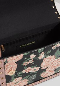 Blommigt mönstrad handväska med svart bakgrund, mjuka rosa rosor, gyllene nitar och ett zip-fodrat innerfack. Märket säger "RIVER ISLAND."