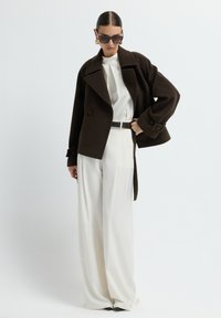 Bruine wollen blendjas met brede revers, gecombineerd met een witte blouse en crème broek met hoge taille. Kenmerkt zich door oversized donkere zonnebril.