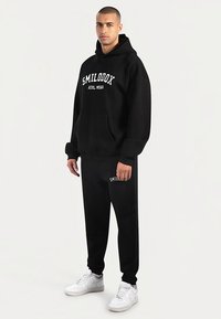 Zwart hoodie en joggingbroek set met witte tekst "SMILODON ATHL WEAR". Voorzien van een voorzak, een ontspannen pasvorm en witte sneakers.