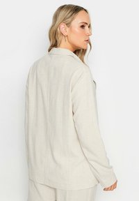Beige pellavablazer, jossa on rento leikkaus, pitkät hihat ja lovellinen kaulus. Kankaalla on tekstuuri ja virtaava siluetti.