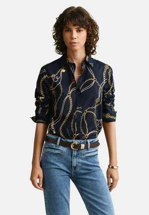 Femme aux cheveux bouclés portant une chemise bleu marine ornée de chaînes dorées, un jean bleu, une ceinture marron et un bracelet doré, posant devant un fond uni.