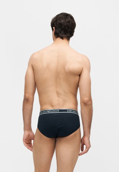 Άνδρας που στέκεται με την πλάτη, φοράει μαύρα εσώρουχα Emporio Armani, με γυμνό κορμό και εμφανή χέρια σε ένα απλό ανοιχτό φόντο.