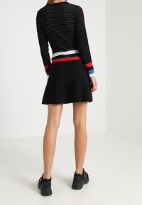 Nainen, jolla on yllään musta hame ja musta pitkähihaine paita punaisilla, valkoisilla ja sinisillä raidoilla, seisoo selkä kameraan päin valkoisessa huoneessa.