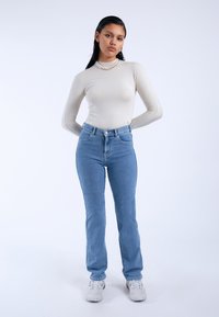 Light beige long-sleeve top paired with high-waisted blue jeans. Model wears white sneakers and layered pearl necklaces, set against a plain background.
