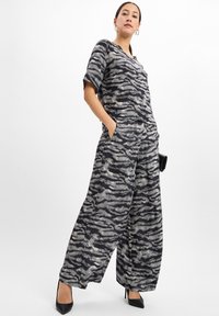 Vrouw die staat in een bijpassende zwarte en grijze dierenprint met wijde broek en korte mouwen top, zwarte puntigehakken, en een kleine clutch vasthoudend.