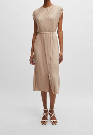 Vrouw draagt een beige mouwloze geplooide midi-jurk met een dunne riem en beige sandalen met hakken en bandjes op een lichte achtergrond.