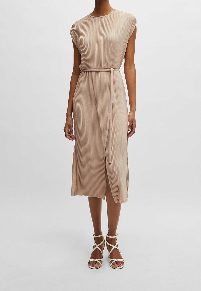 Femme portant une robe midi plissée beige sans manches avec une fine ceinture et des sandales à talons beige à lanières sur un fond clair.