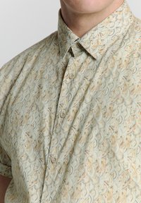 Kurzarmhemd aus hellgrünem Stoff mit einem wiederkehrenden Wellenmuster in Beige und Braun, ausgestattet mit einem Kragen zum Knöpfen und Vorderknöpfen.