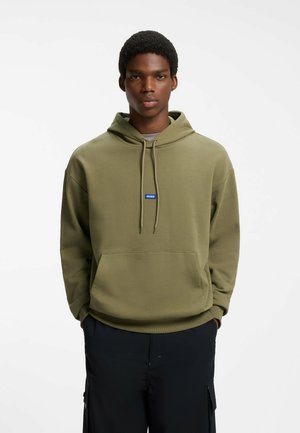 Jonge man draagt een olijfgroene hoodie met voorzak en zwarte broek, staand tegen een witte achtergrond.