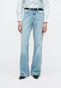 Jasnoniebieskie dzwony z denimu, z wysokim stanem, pięcioma kieszeniami i subtelnym przetarciem. Stylizowane z czarnym paskiem i szpiczastymi szpilkami.