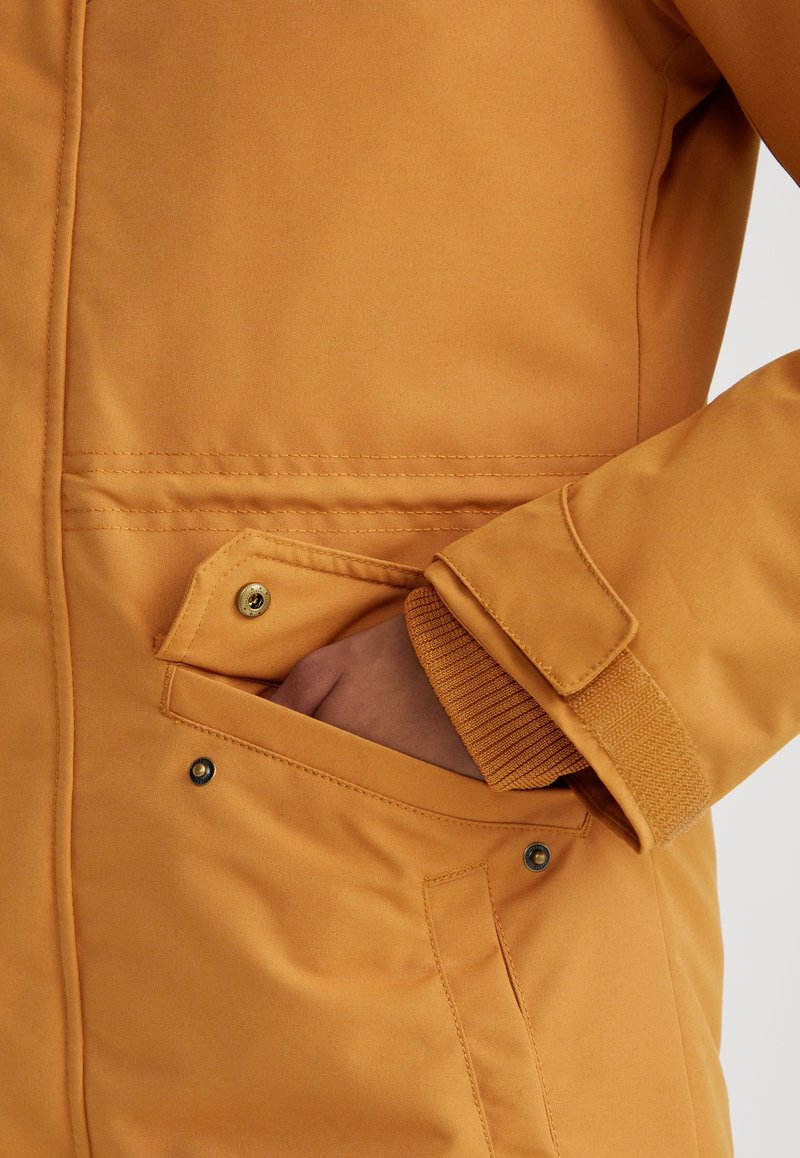 Chaqueta de color mostaza con una textura suave, que cuenta con un bolsillo lateral con solapa y cierre a presión, y puños acanalados para un mejor ajuste.