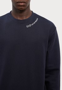Granatowa bluza crewneck wykonana z miękkiego materiału, cechująca się teksturowanym wykończeniem. Logo wyszyte na biało na lewym ramieniu.