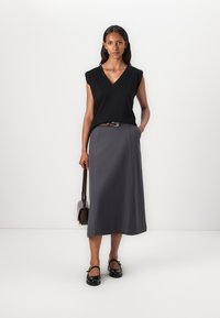 Schwarzes V-Ausschnitt-Oberteil mit kurzen Ärmeln, kombiniert mit einem langen grauen Rock und einem braunen Gürtel. Tragen mit schwarzen flachen Schuhen und einer braunen Handtasche.