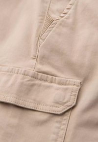 Primer plano de tela beige con costuras detalladas en la solapa de un bolsillo y la costura de una prenda.