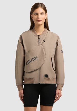 Woman wearing a beige zip-up bomber jacket, a matching crossbody bag with "KHUJO" text, and black biker shorts, standing against a neutral background.