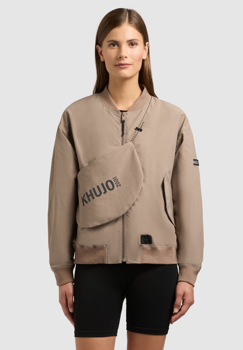 Femme portant une veste bomber beige à fermeture éclair, un sac bandoulière assorti avec l'inscription "KHUJO", et un short de cycliste noir, debout devant un fond neutre.