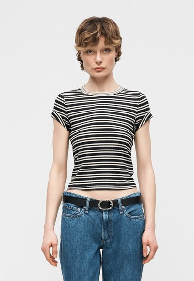 Νεαρός με κοντά κυματιστά μαλλιά, που φοράει μαύρο και άσπρο ριγέ κοντομάνικο πουκάμισο και μπλε τζιν με μαύρη ζώνη.