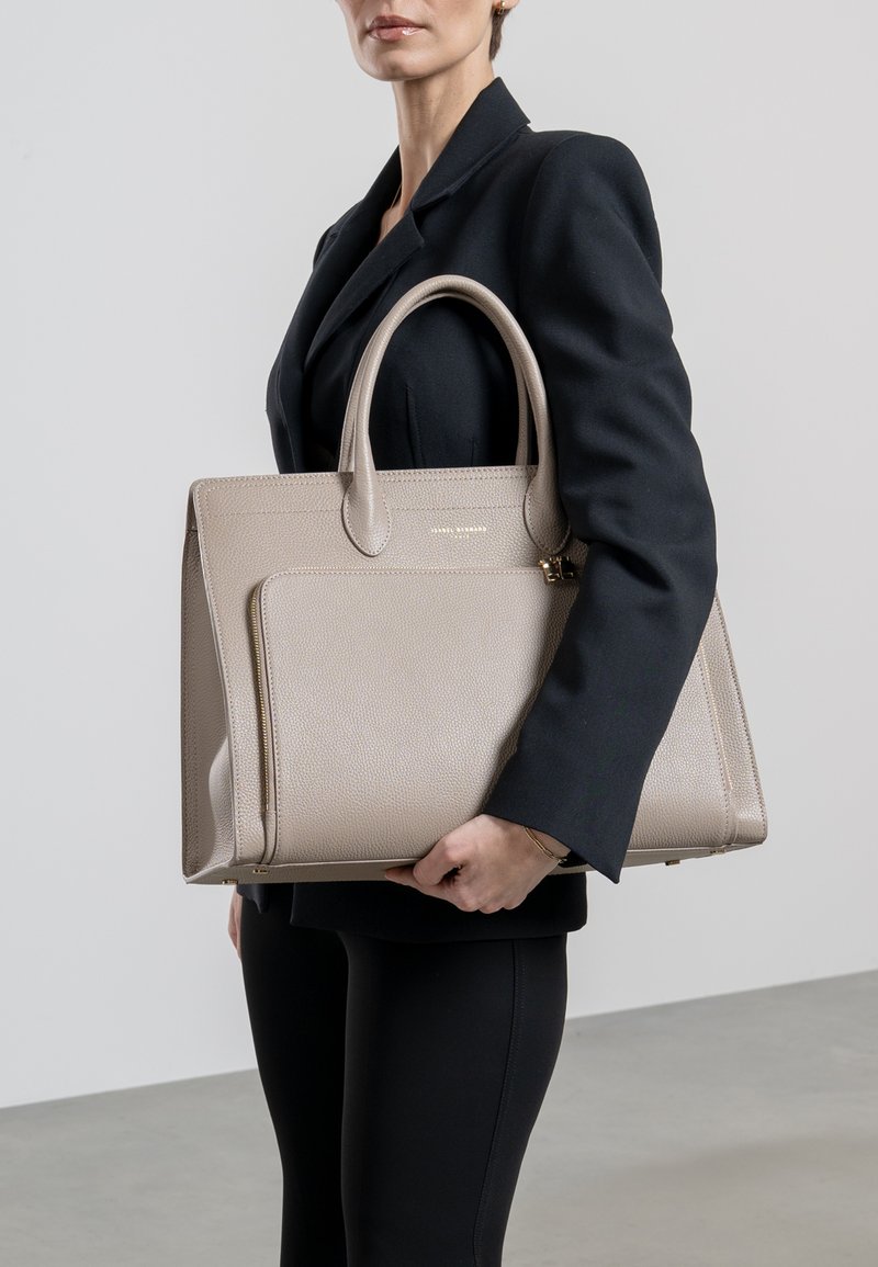 Beige leather tote bag with structured shape, dual top handles, and a front zip pocket. Textured finish with gold hardware accents.