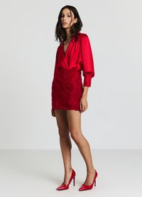 Blusa rossa con scollo a V profondo e maniche a sbuffo, abbinata a una mini gonna rossa testurizzata con applicazioni di rose. Tacchi alti rossi completano il look.