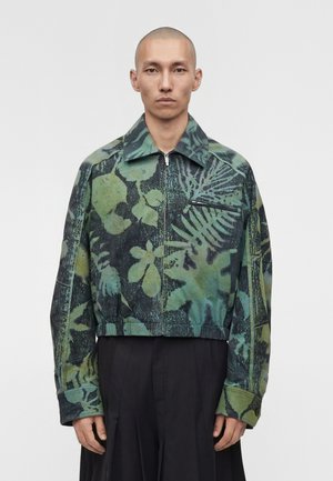 Άνδρας με ξυρισμένο κεφάλι που φοράει πράσινο και μαύρο μπουφάν με φερμουάρ και φλοράλ σχέδιο και μαύρο παντελόνι με φαρδιά πόδια, στέκεται σε λευκό φόντο.