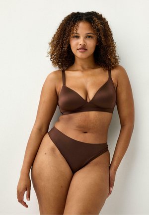 Kvinde med krøllet hår iført en brun bh og matchende højskårede trusser, stående mod en ensfarvet lys baggrund.