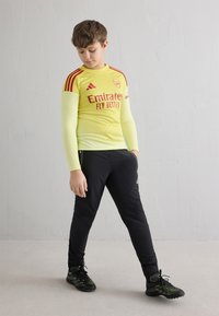 Žltý športový dres s dlhým rukávom, červenými akcentmi a logom, doplnený čiernymi teplákmi a čiernymi športovými topánkami so zelenými detailmi.