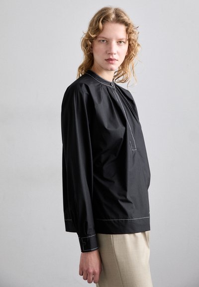Blusa negra de manga larga con un cuello alto, acentos de costura blanca y un corte holgado. Tejido suave, detalles de botones en los puños y el escote.
