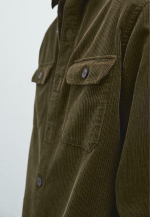 Camicia in corduroy verde oliva con due tasche anteriori con bottone, texture a righe verticali e colletto. I bottoni sono di colore marrone scuro.