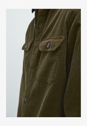 Olivgrünes Cordhemd mit zwei geknöpften Brusttaschen, vertikaler Rippstruktur und Kragen. Die Knöpfe sind dunkelbraun.