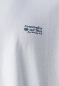 Hvidt stof med lille broderet tekst "Abercrombie and Fitch NYC New York City, N.Y." med mørk tråd.